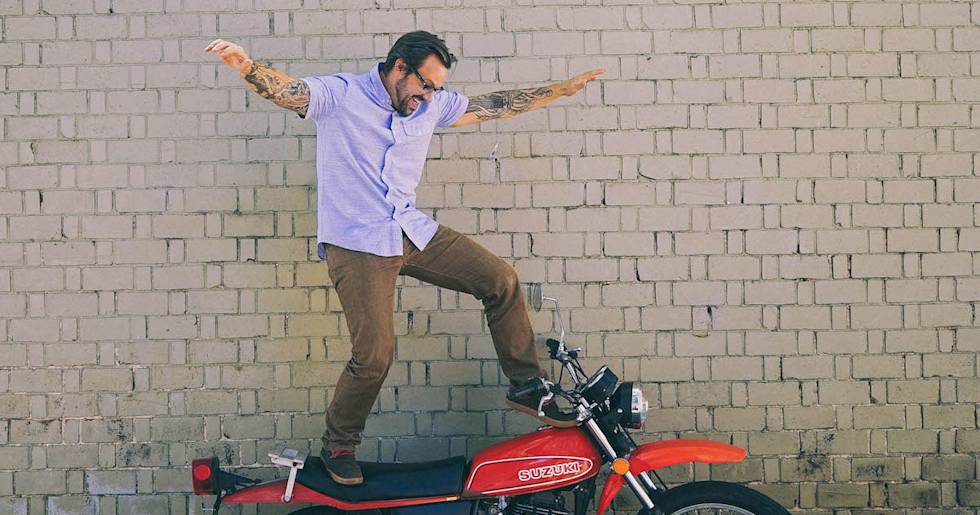 Energetic person with tattoos balancing playfully on a red Suzuki motorcycle against a brick wall, arms spread wide in celebration