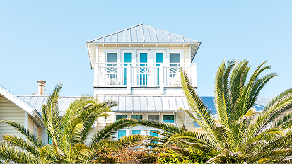 Row of white coastal homes with metal roofs seen through tropical palm fronds against bright blue sky