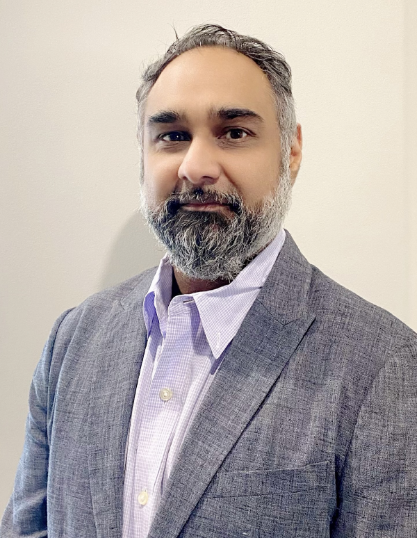 Professional headshot of a person with a salt-and-pepper beard wearing a gray suit jacket and light purple checkered shirt