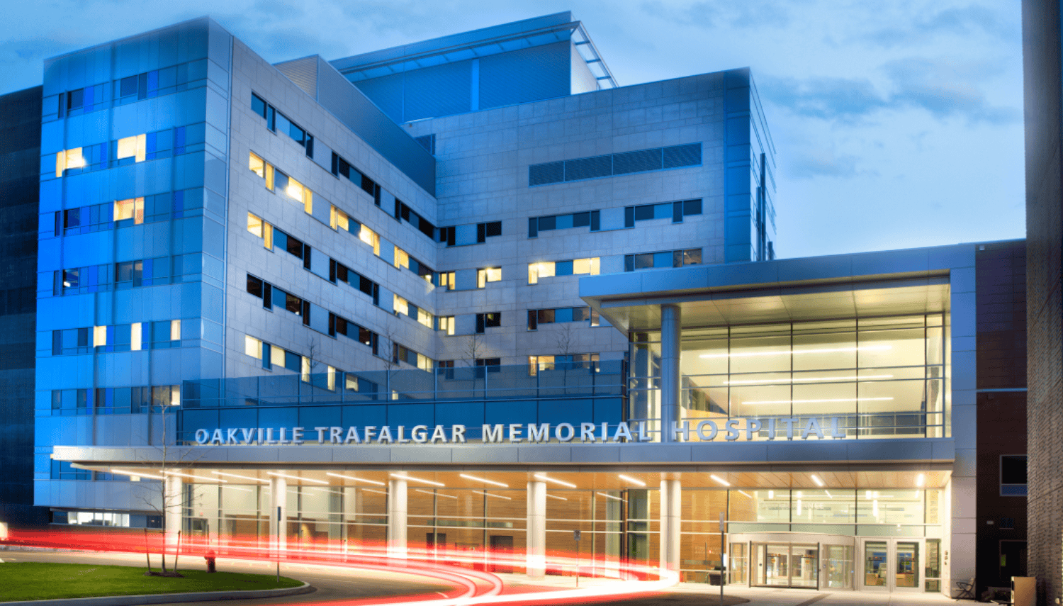 View of Oakville Trafalgar Memorial Hospital at dusk. The main entrance is well-lit. Cars drive by, leaving trails of light.