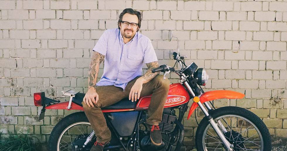 Tattooed motorcyclist in lavender shirt and glasses sits on vintage red Suzuki bike against white brick wall