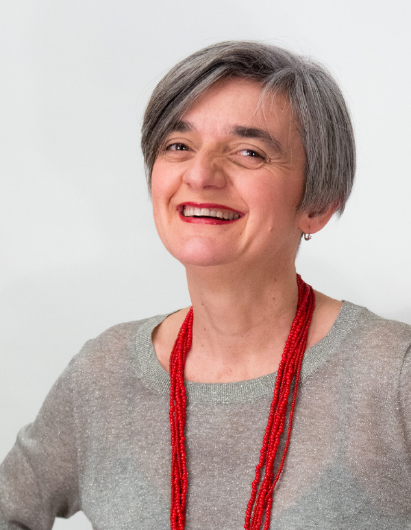 A person with short gray hair wearing a gray sweater and vibrant red beaded necklace smiles warmly against a white background