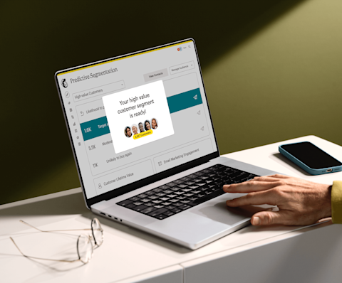 Image of a person sitting at a desk, looking at a laptop screen displaying Mailchimp’s Predictive Segmentation feature. The visual shows a display indicating that the user’s high value customer segment is ready to view. 