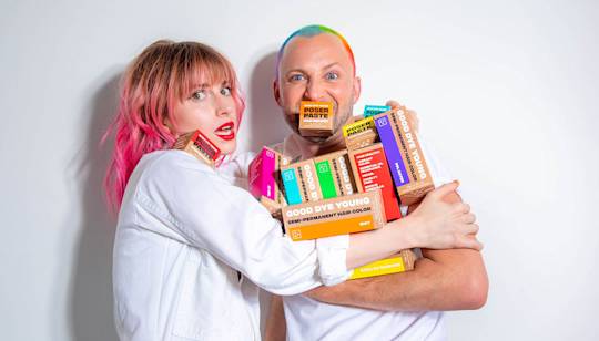 Two people in white shirts holding colorful Good Dye Young hair product boxes, one with pink hair and one with rainbow-dyed mohawk