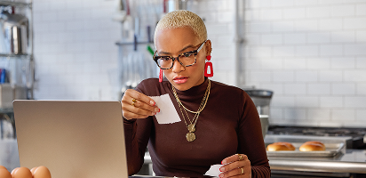 Professional with short blonde hair and glasses working at laptop while holding coffee cup in modern kitchen setting