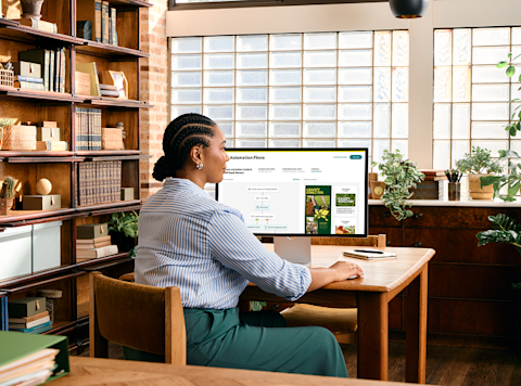 Professional working at wooden desk in library-like setting with bookshelves and plants, viewing computer screen in natural light