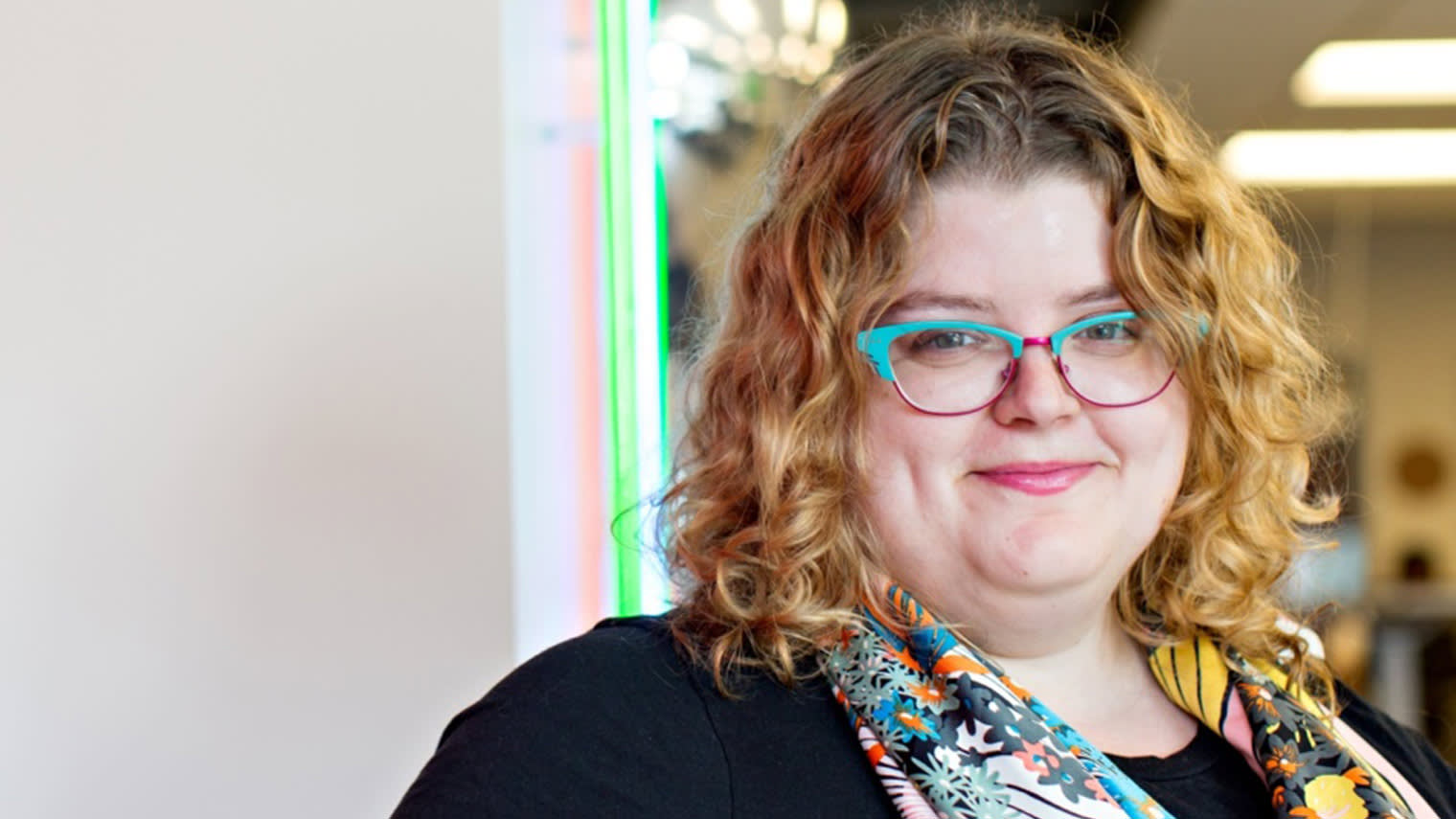 A smiling person with curly blonde hair wearing bright turquoise and red glasses and a black top with colorful floral pattern