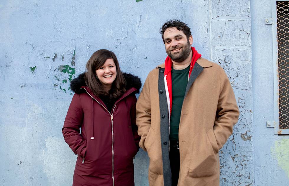 Ric and Morgan from the Adhoc Presents team stand smiling against a light blue wall. 