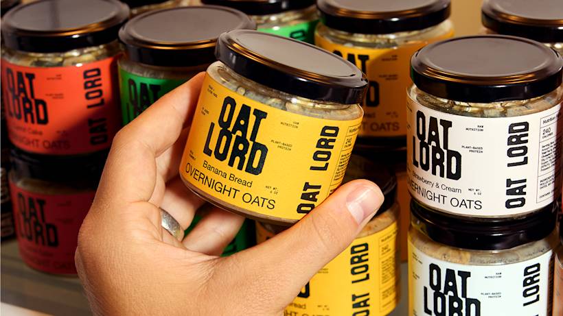 Hand reaching for jar of Oat Lord banana bread overnight oats among rows of colorful protein oat jars on shelf