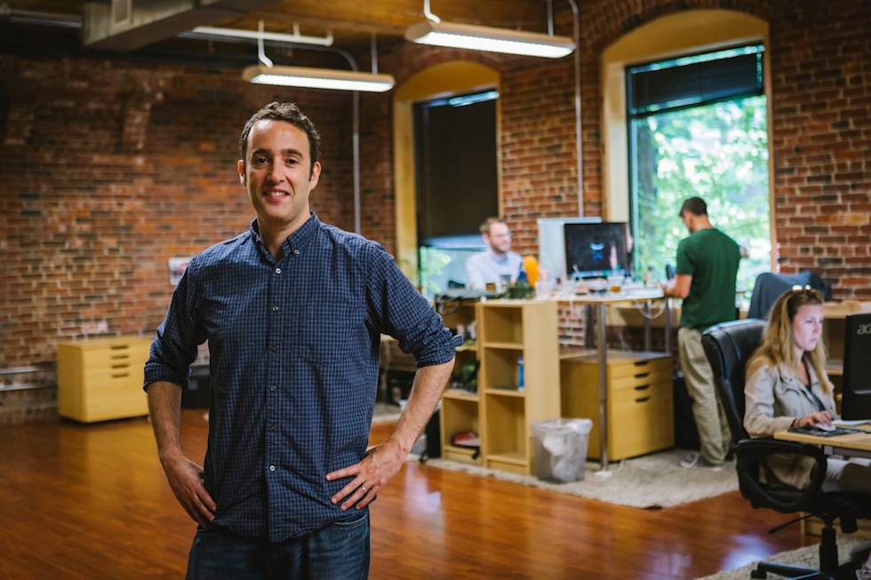Professional standing in modern office space with exposed brick walls, wooden floors, and team members working in background
