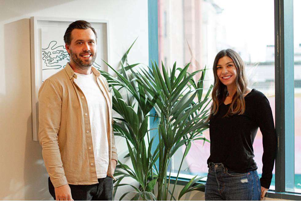 Two professionals standing in a bright office space with a large palm plant between them, near a window with modern decor
