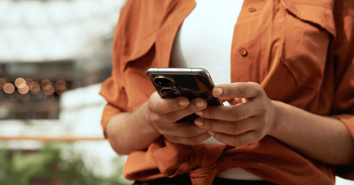 Close-up of a person's hand holding a smartphone with dual cameras, wearing an orange shirt over a white t-shirt.