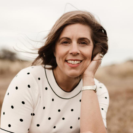 A person wearing a white polka dot top smiles warmly at the camera while their dark hair blows in the wind against a blurred outdoor background