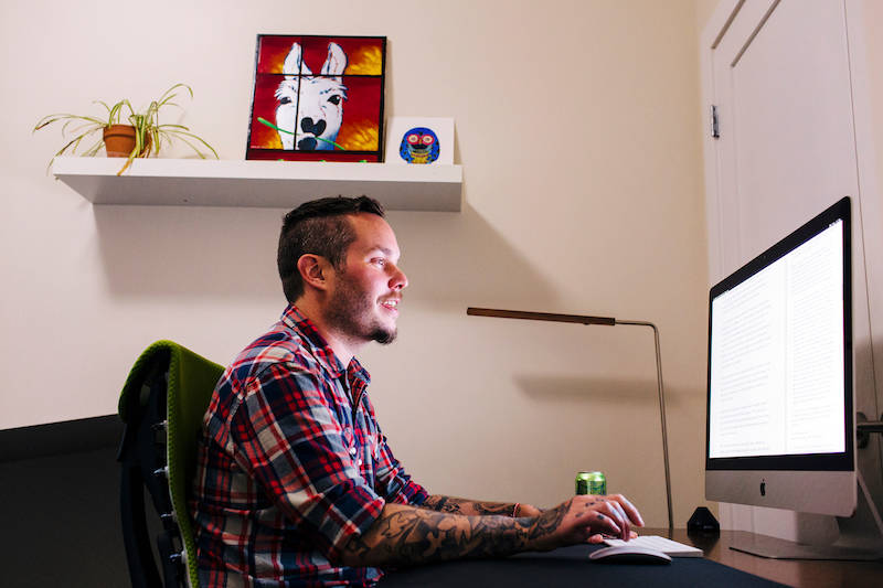 Person in plaid shirt working at desk with iMac computer, illuminated by desk lamp, with art and plant on wall shelf above