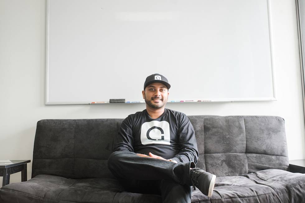A person in a black baseball cap and long-sleeved shirt with a logo sits casually on a gray sectional couch in a bright, minimalist room.