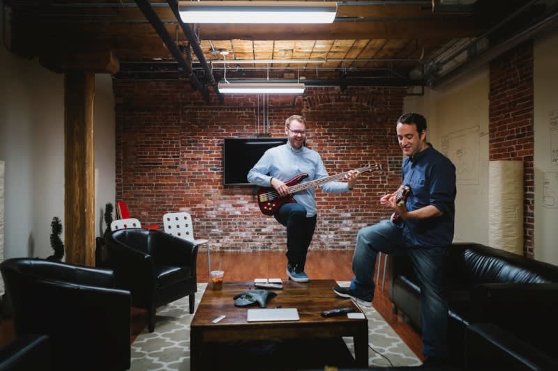Two musicians playing guitars in a casual brick-walled lounge area with black leather couches and a wooden coffee table