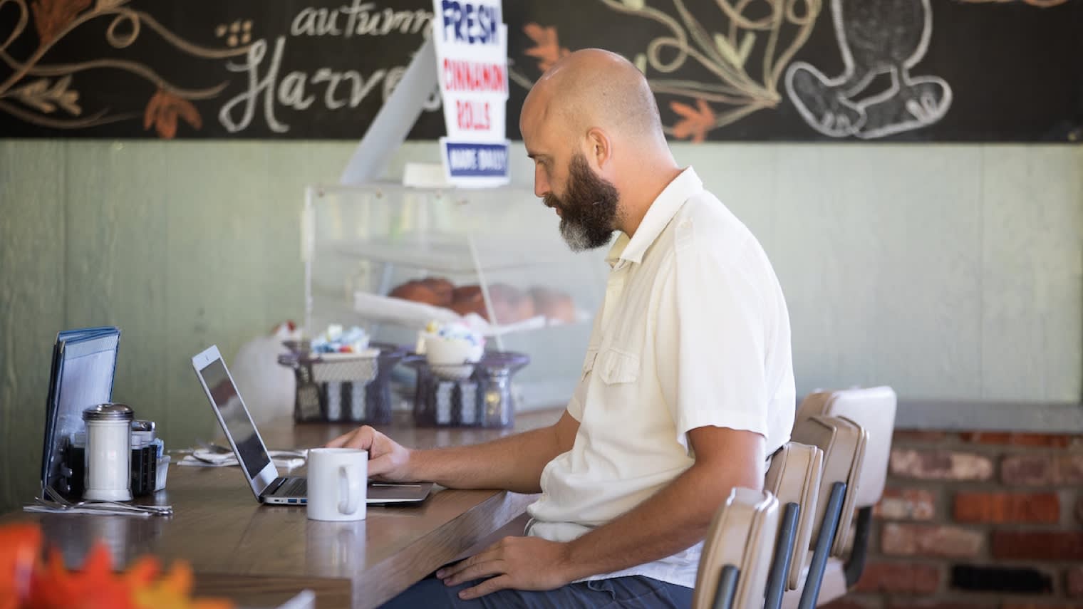 Bearded man in white shirt working on laptop at cafe counter with coffee cup nearby, autumn-themed chalkboard art visible above