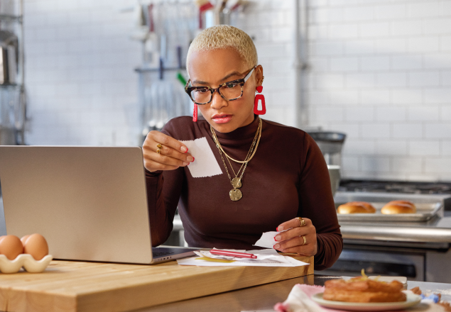Professional working at kitchen counter with laptop, wearing brown turtleneck and glasses while reviewing documents