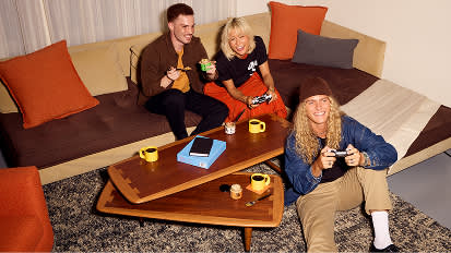A group of people relaxing on a gray couch with orange pillows, sharing drinks and snacks on wooden serving trays