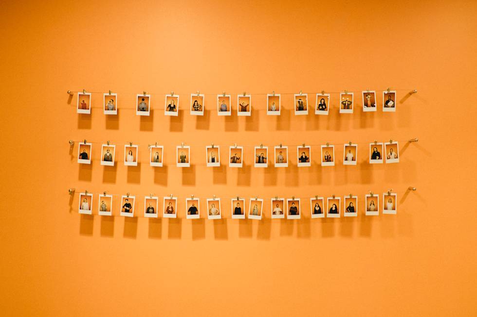 Three rows of instant photos hung on strings against an orange wall, displaying portrait photographs arranged in a gallery style