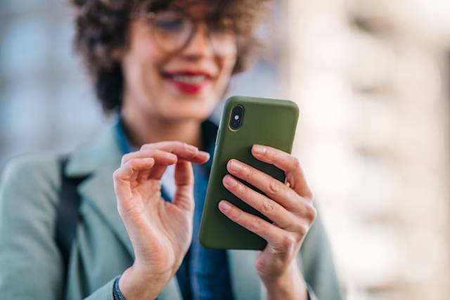Close-up of hands holding a green smartphone case, with a smiling person wearing glasses and red lipstick in the background