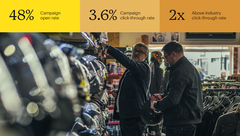 Two men are browsing helmets in a motorcycle shop, both wearing dark jackets and eyeglasses. One man is reaching toward a shelf with a variety of motorcycle helmets. The other man is holding a black helmet.