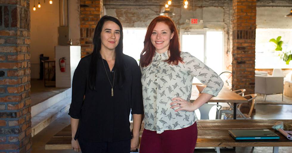 Two professionals standing in modern office space with exposed brick walls and pendant lighting, one wearing black, one in patterned blouse