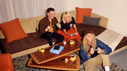 Three friends relaxing in living room with coffee and snacks, sitting on beige couch with orange pillows and wooden coffee table