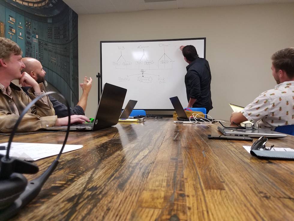 Team meeting in conference room with whiteboard diagram, laptops on wooden table, and industrial-style wall mural in background