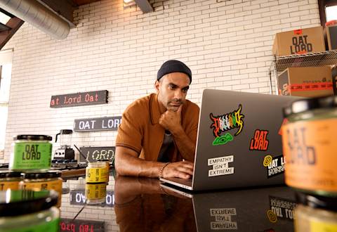 Entrepreneur working on laptop with Oat Lord branded products and signage displayed in background against white brick wall